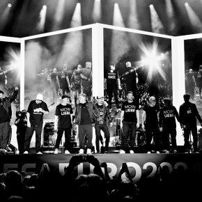 Die Fantastischen Vier und weitere Personen stehen nebeneinander am Bühnenrand. Im Hintergrund sind die Personen mehrfach von hinten auf Leinwänden zu sehen. Im Vordergrund ist schemenhaft das Publikum zu erahnen.