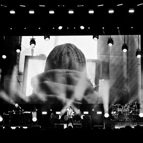 Das aus der Ferne frontal aufgenommene Schwarz-Weiß-Foto zeigt die Musiker und den Bühnenaufbau eines Konzerts der Band Porcupine Tree. Im Hintergrund befindet sich eine große Leinwand, über die offenbar ein Video gezeigt wird. Zu sehen ist ein Mnesch von hinten mit Kapuzenpullover und Strickmütze.