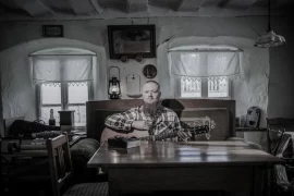 Nico Schwappacher (Zwischenlichten) sitzt mit einer Akustikgitarre am Tisch in einer ländlichen Stube.